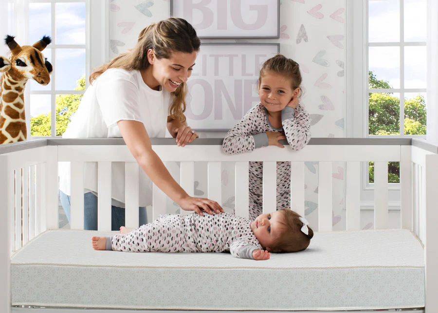 Baby on crib mattress with mom and big sister looking on along with a stuffed giraff
