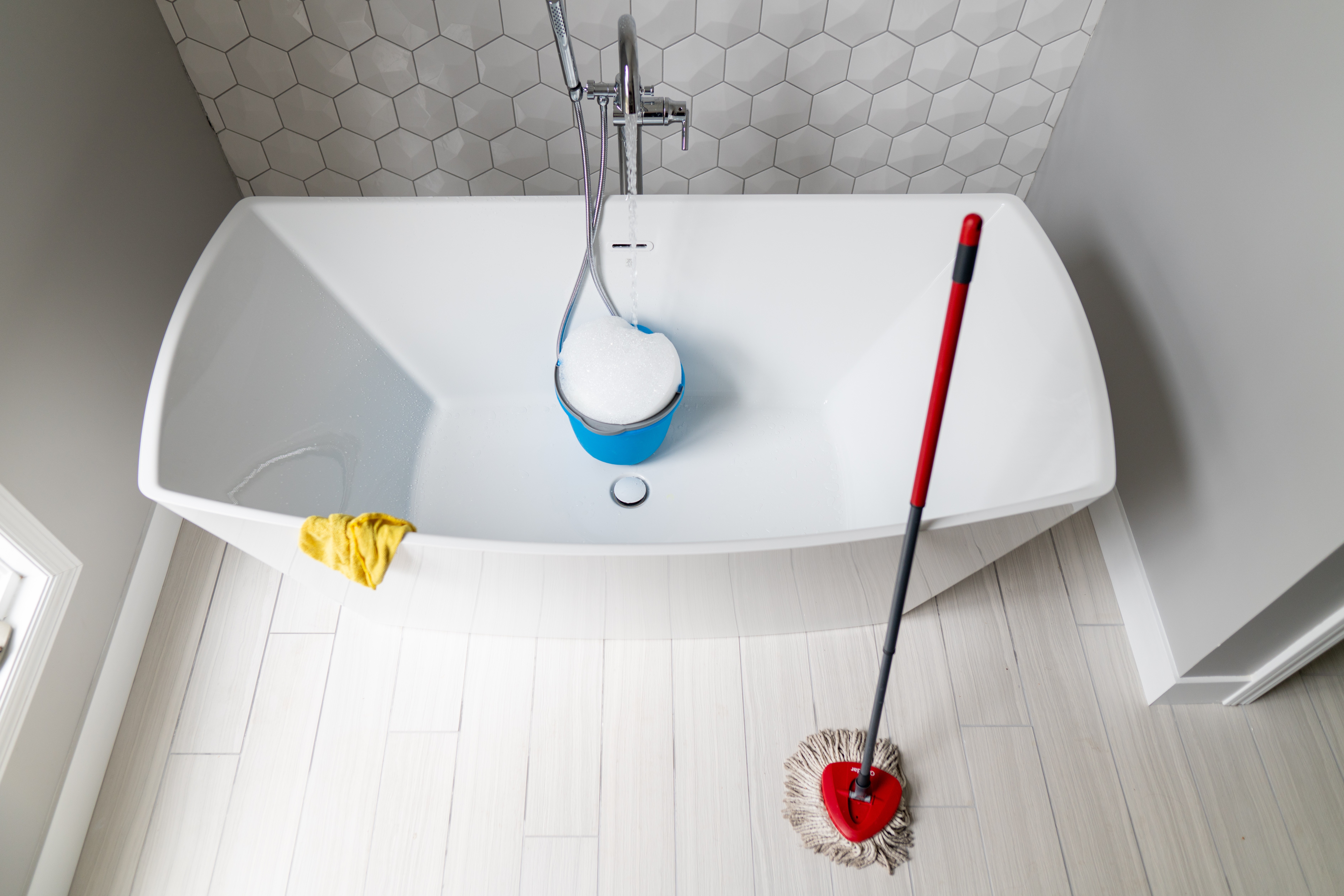 Mop leaning against tube with a sudsy bucket in the tub and yellow rubber gloves hanging over the side